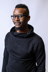 May include: A person wearing a black hooded sweatshirt with a high neckline and a small, stitched design on the front.
