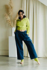 May include: A woman wearing a lime green cropped zip-up sweatshirt and dark blue wide-leg jeans. The outfit includes matching lime green socks. The background features a white vase with dried plants and a white curtain.