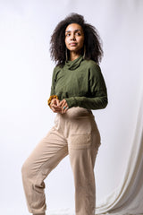 May include: A woman wearing a green long-sleeved shirt with a high neck and beige pants. She is standing in front of a white background.