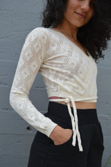 May include: A cream-colored, long-sleeved, wrap-style sweater with a diamond-shaped pattern. The sweater has a tie at the waist.