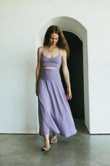May include: A two-piece lavender outfit consisting of a crop top and a flowing maxi skirt. The top has thin straps and a ruched bust. The skirt is high-waisted and falls to the ankles. The outfit is made of a soft, flowing fabric.