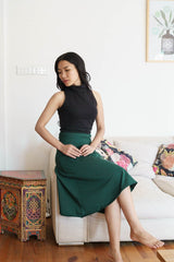 May include: A woman wearing a black mock neck sleeveless top and a dark green skirt. She is sitting on a white couch with patterned pillows.